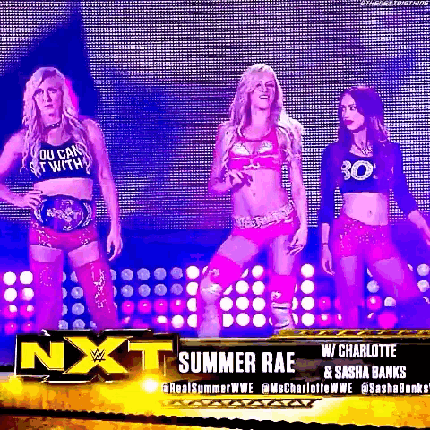 three women are standing on a stage in front of a sign that says nxt summer rae .