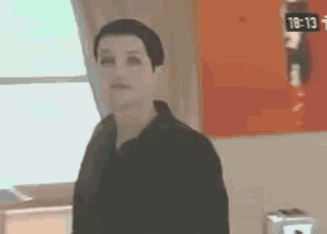 a man in a black shirt is standing in front of a window with a clock on the wall that says 18:13