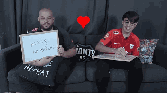 two men are sitting on a couch holding signs that say kebab hamburguesa and repeat