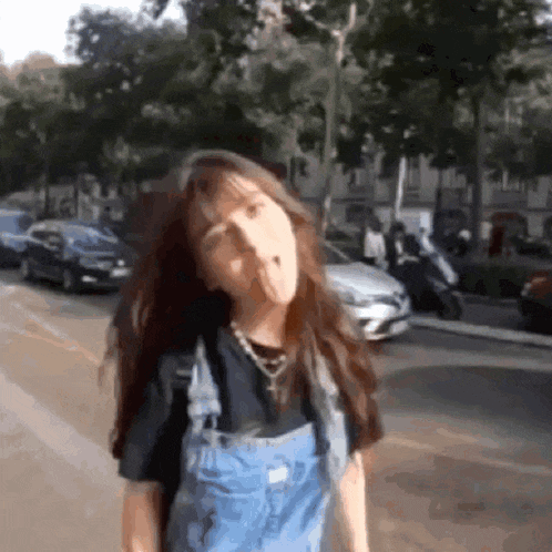 a girl wearing overalls and a black shirt is standing on a street making a funny face .