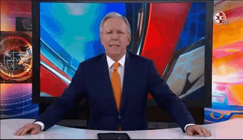 a man in a suit and orange tie is standing in front of a television