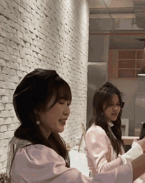 two women are sitting in front of a white brick wall and smiling
