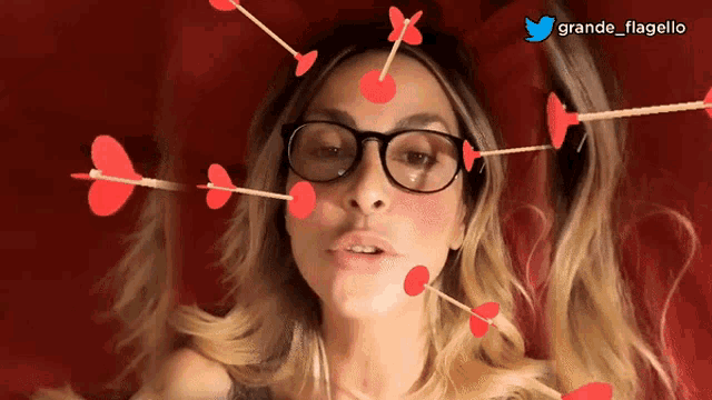 a woman wearing glasses is surrounded by red hearts and arrows with the twitter handle grande flagello