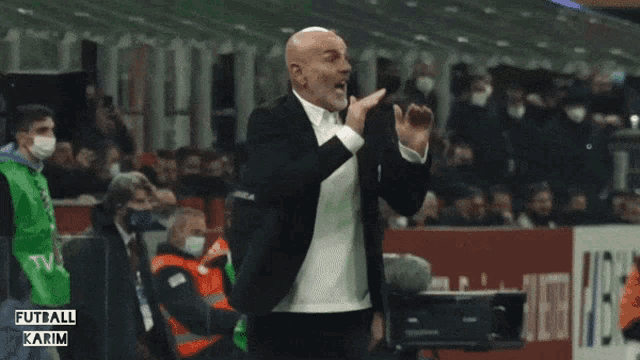 a man in a suit stands on a soccer field with the words futball karim on the bottom left