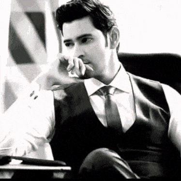 a black and white photo of a man in a suit and tie sitting in a chair .