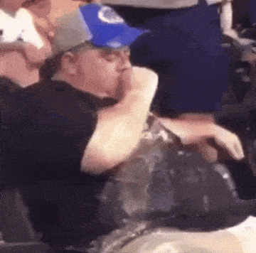 a man in a blue hat is sitting in a stadium holding a bottle .