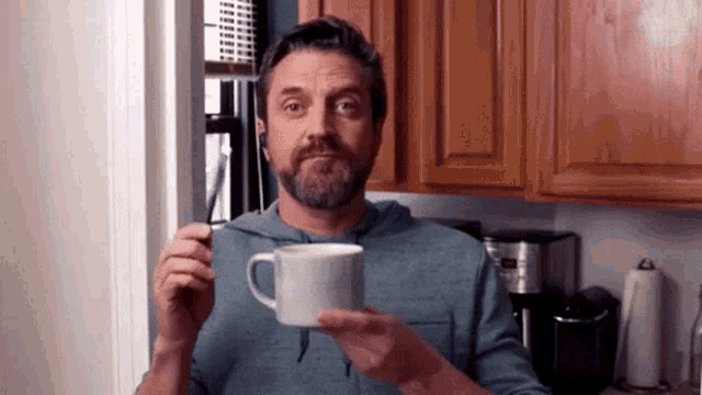 a man with a beard is holding a cup of coffee in his hand in a kitchen .