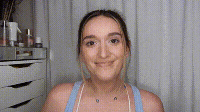 a woman wearing a blue tank top and a necklace is smiling in front of a white curtain .