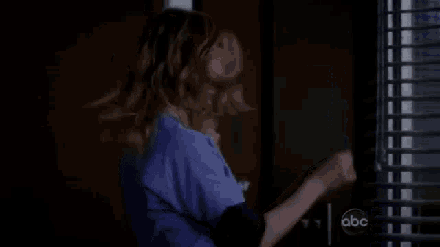 a woman in a blue scrub top is opening a window with blinds on it .