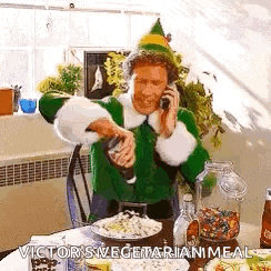 a man dressed as a christmas elf is talking on a cell phone while sitting at a table with food .