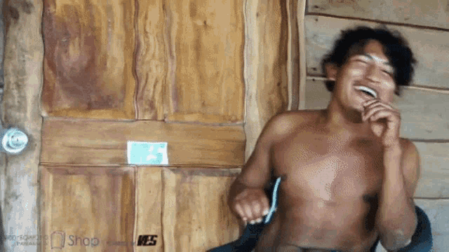 a shirtless man is laughing in front of a wooden door with a green sticker on it that says shop