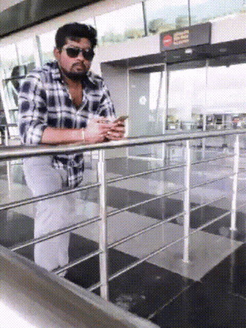 a man wearing sunglasses and a plaid shirt is standing on a railing looking at his phone