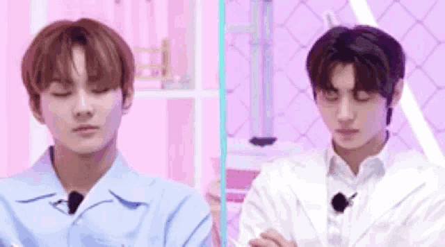 two young men are sitting next to each other in front of a pink wall . one of the men is wearing a blue shirt .