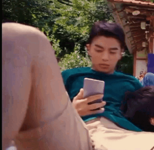a man in a blue shirt is sitting on the ground holding a cell phone in his hand .