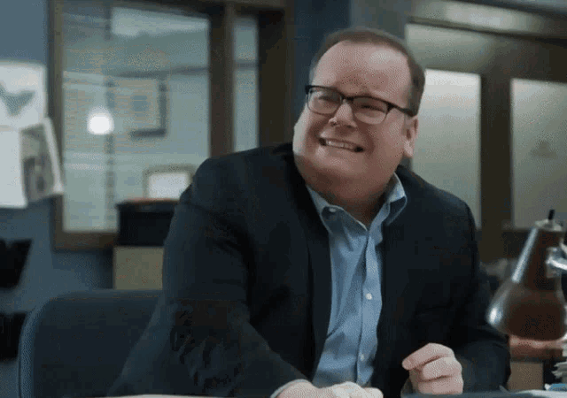 a man in a suit and glasses sits at a desk with his mouth open