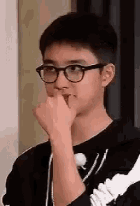 a young man wearing glasses and a black shirt is holding his hand to his mouth .