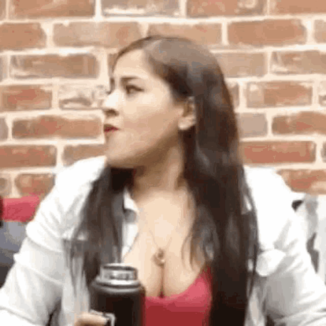 a woman is sitting in front of a brick wall holding a thermos and making a funny face .