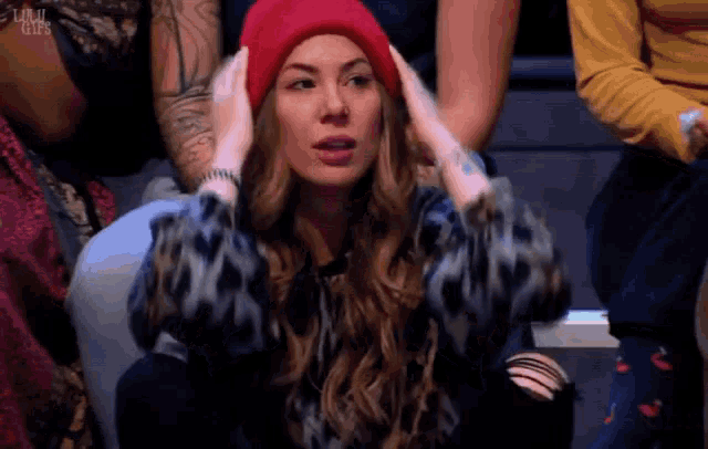 a woman wearing a red beanie holds her hands to her ears while sitting in a crowd