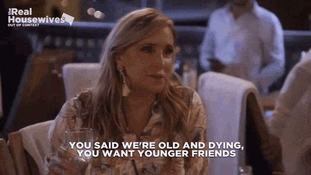 a woman is sitting at a table with a glass of wine and says " you said we 're old and dying "