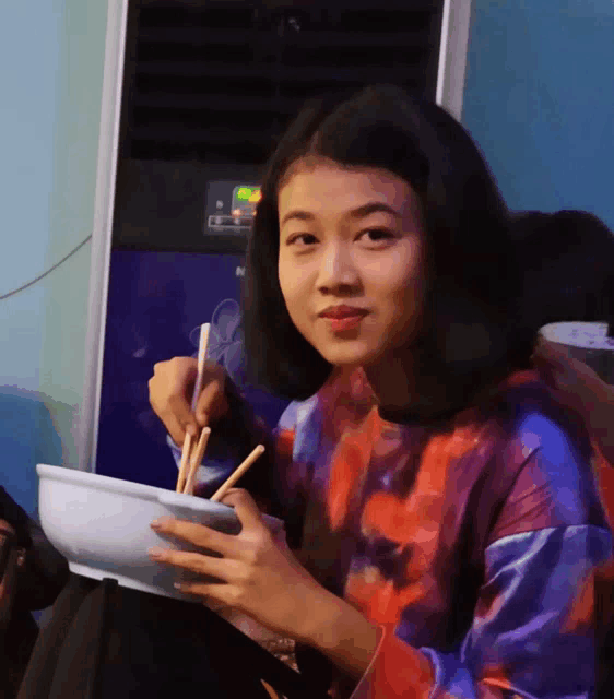 a woman eating a bowl of food with chopsticks in front of an air conditioner that says n on it