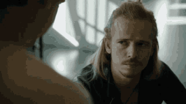 a man with long hair and a mustache is looking at another man 's back .