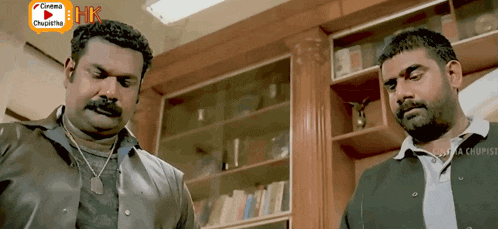 two men are standing in front of a bookshelf with a bhk logo on the bottom