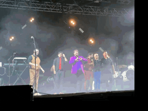 a group of men standing on a stage with a man in a purple jacket playing a violin