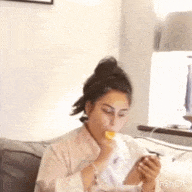 a woman is sitting on a couch eating a piece of food while looking at her phone .