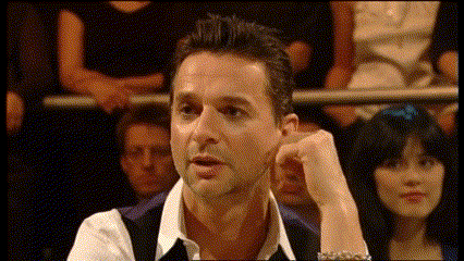 a man in a white shirt and black vest holds his hand to his ear in front of a crowd of people