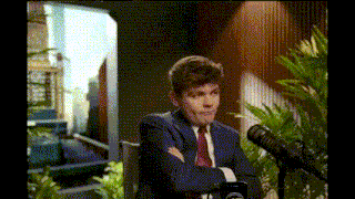 a man in a suit and tie is sitting in front of a microphone with his hands folded .