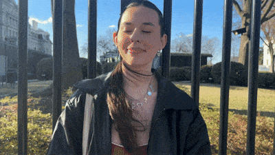 a woman wearing a black jacket and a necklace stands in front of a fence with her eyes closed