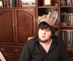 a man with a beard wearing a hat is sitting in front of a bookshelf .