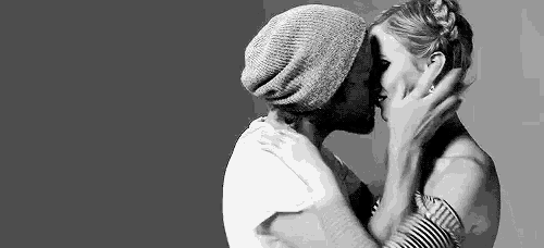a man and a woman are kissing in a black and white photo . the woman is wearing a hat .