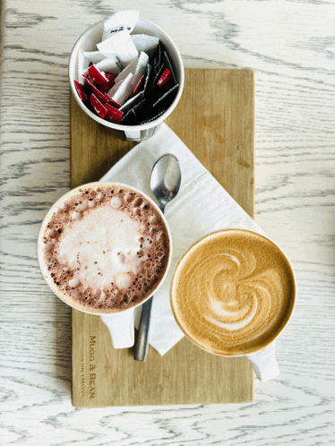 two cups of coffee are on a wooden cutting board that says ' maggi & bean ' on it