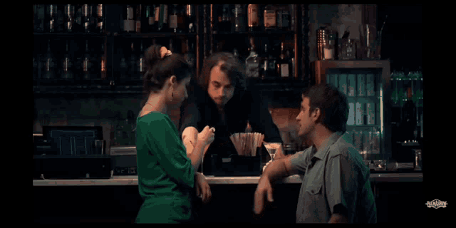 a woman in a green dress sits at a bar talking to a man and a bartender