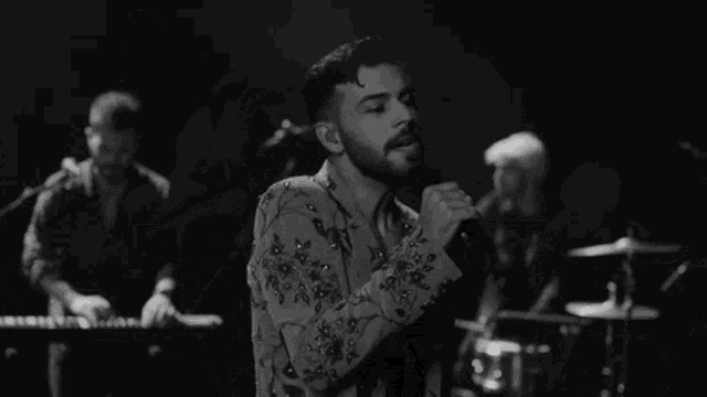 a black and white photo of a man singing into a microphone while playing drums .