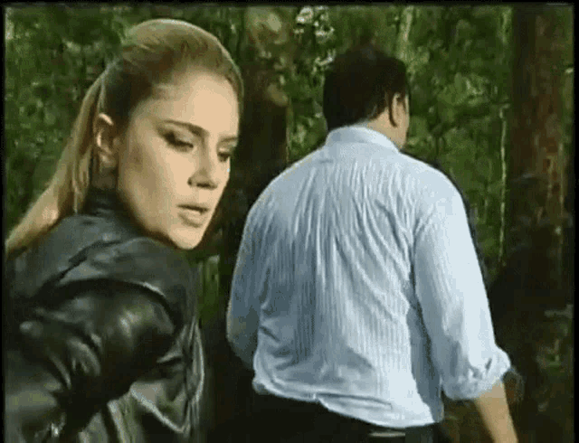 a man and a woman are standing in the woods . the woman is wearing a black leather jacket .