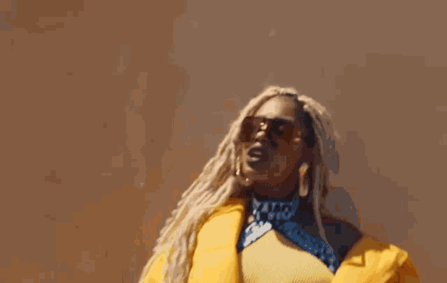 a woman wearing a yellow jacket and sunglasses is standing in front of a brown wall .