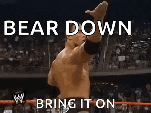 a wrestler is standing in a wrestling ring with his arms in the air and the words `` bear down bring it on '' .