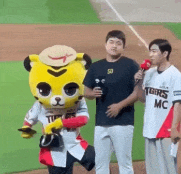 a mascot is standing next to two men on a baseball field holding a microphone .