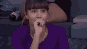 a woman in a purple shirt is sitting on a couch covering her mouth with her hand .