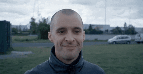 a man with a shaved head is smiling in a field with a van in the background