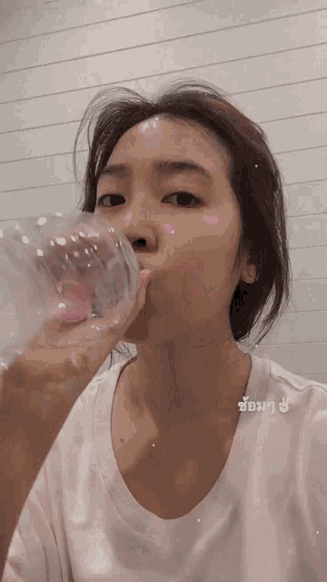 a woman in a white shirt drinks from a clear plastic bottle in front of a white wall
