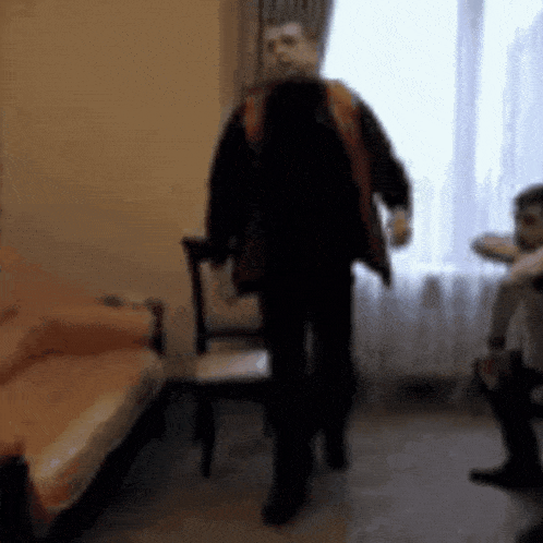 a man in a black jacket is standing in a living room next to a couch and chair .