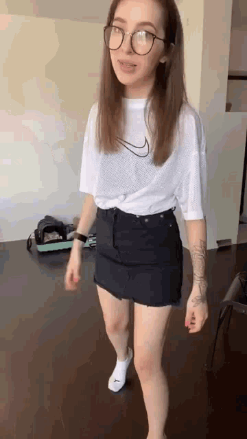 a woman wearing glasses , a white t-shirt and a black skirt is standing on a wooden floor .
