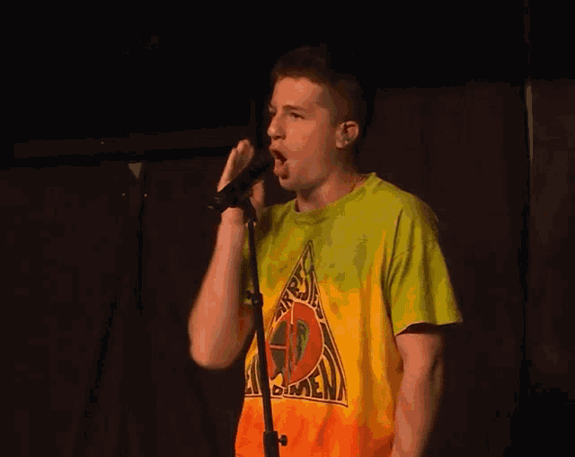 a young man singing into a microphone wearing a tie dye shirt that says ' reggae ' on the front