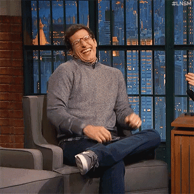a man is sitting in a chair laughing while wearing glasses and a sweater .