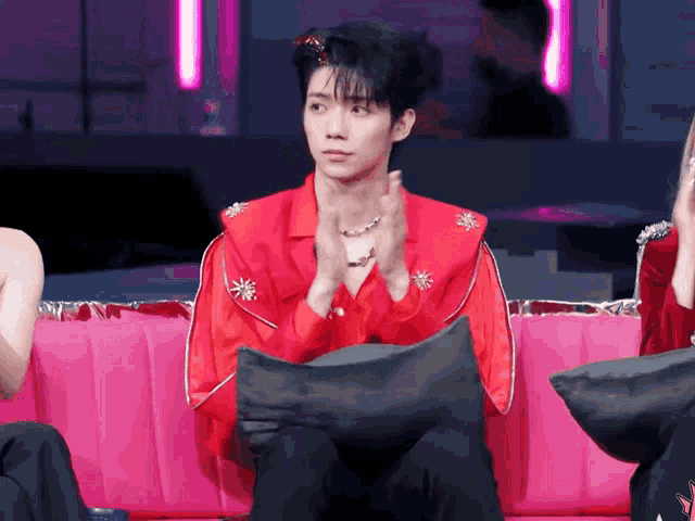 a man in a red jacket sits on a pink couch clapping