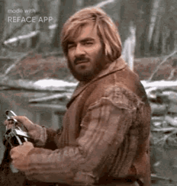 a man with a beard and long blonde hair is standing in the woods holding a gun .