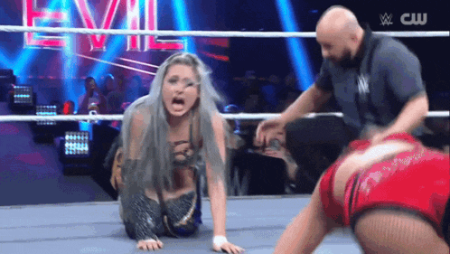 a woman is kneeling on the ground in a wrestling ring while a referee stands behind her .